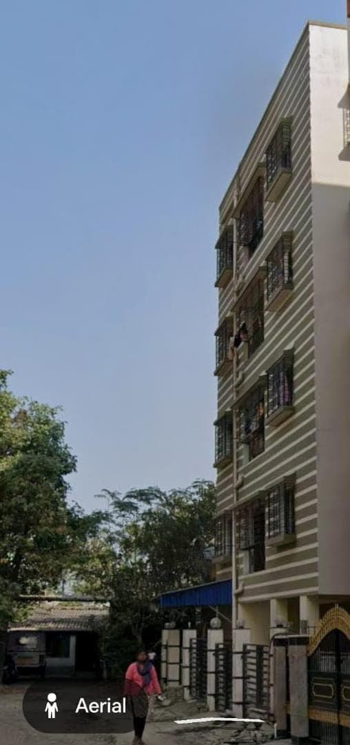 The same building seen along the street — its full height above the boundary wall and entrance gate, with the neighbouring plot and trees to the left